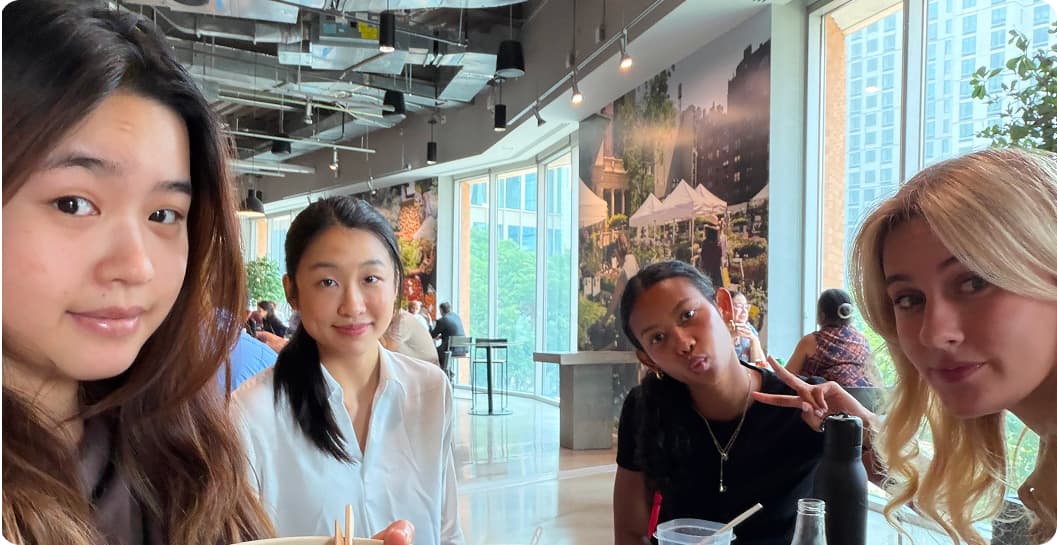 Four interns posing for a selfie over lunch, with a mural of a city park and skyline in the windows behind them.