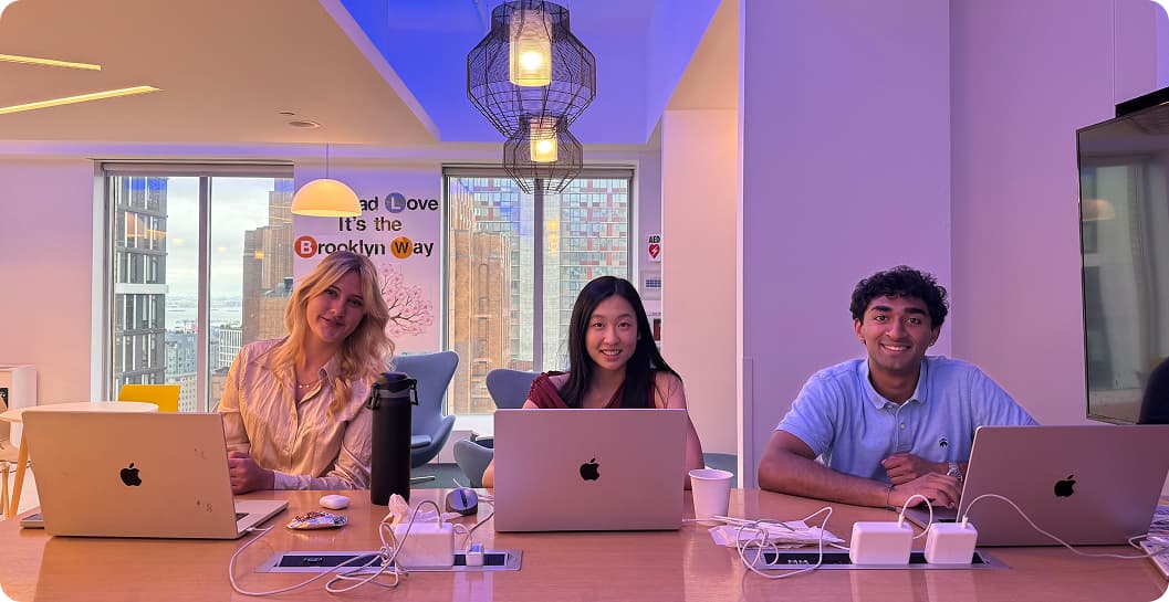 Three interns smiling at a shared desk with open laptops, in a lounge with a “We'd Love It's the Brooklyn Way” sign in the background.