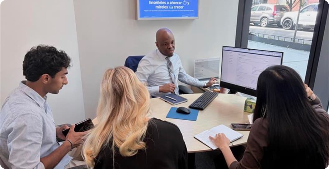 Four people gathered around a meeting-room table, one banker explaining a dashboard on a desktop monitor while two others take notes.